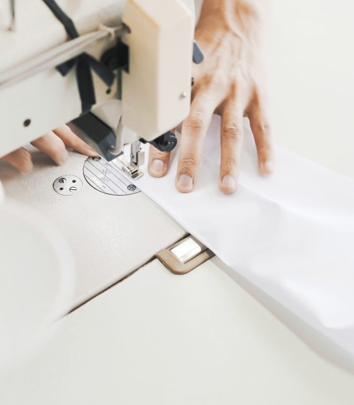 Female hands with white sewing machine, top view shot. Spool of thread blurred on foreground, female hands sewing white cloth on white table, bright modern atelier studio