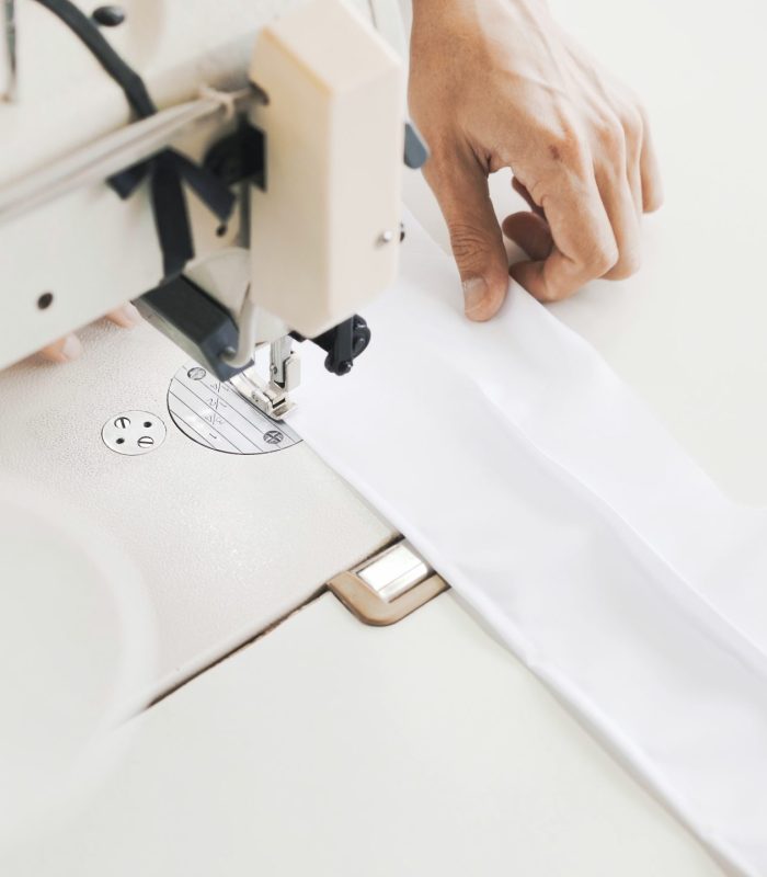 Female hands sewing white sewing machine, top view shot. Spool of thread blurred on foreground, female hands sewing white cloth on white table, bright modern atelier studio
